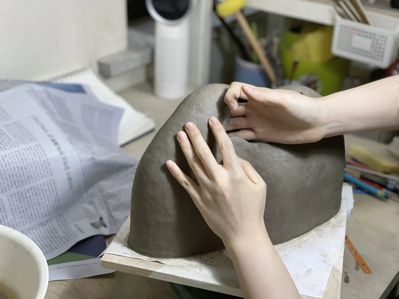 Hands shaping a large clay form on a studio table